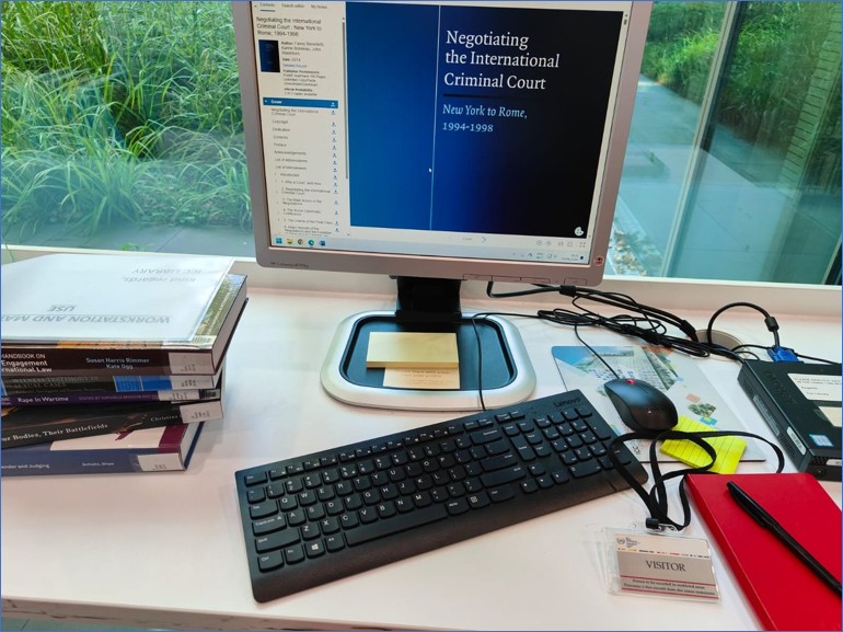 The researcher’s designated desk at the ICC Library, under a surveillance camera. On the desk are books—including Rape in Wartime; Our Bodies, Their Battlefields; and compilations of feminist civil society reporting from the Rome Statute negotiations—and a visitor badge. (Photo by the author)